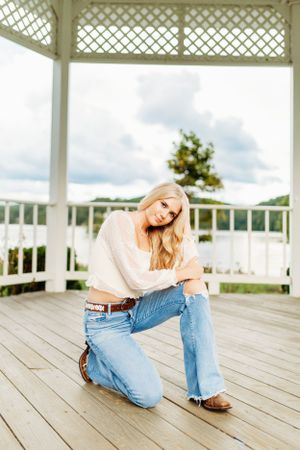 Blonde teen in white top and distressed jeans kneeling in a white gazebo for senior portraits.