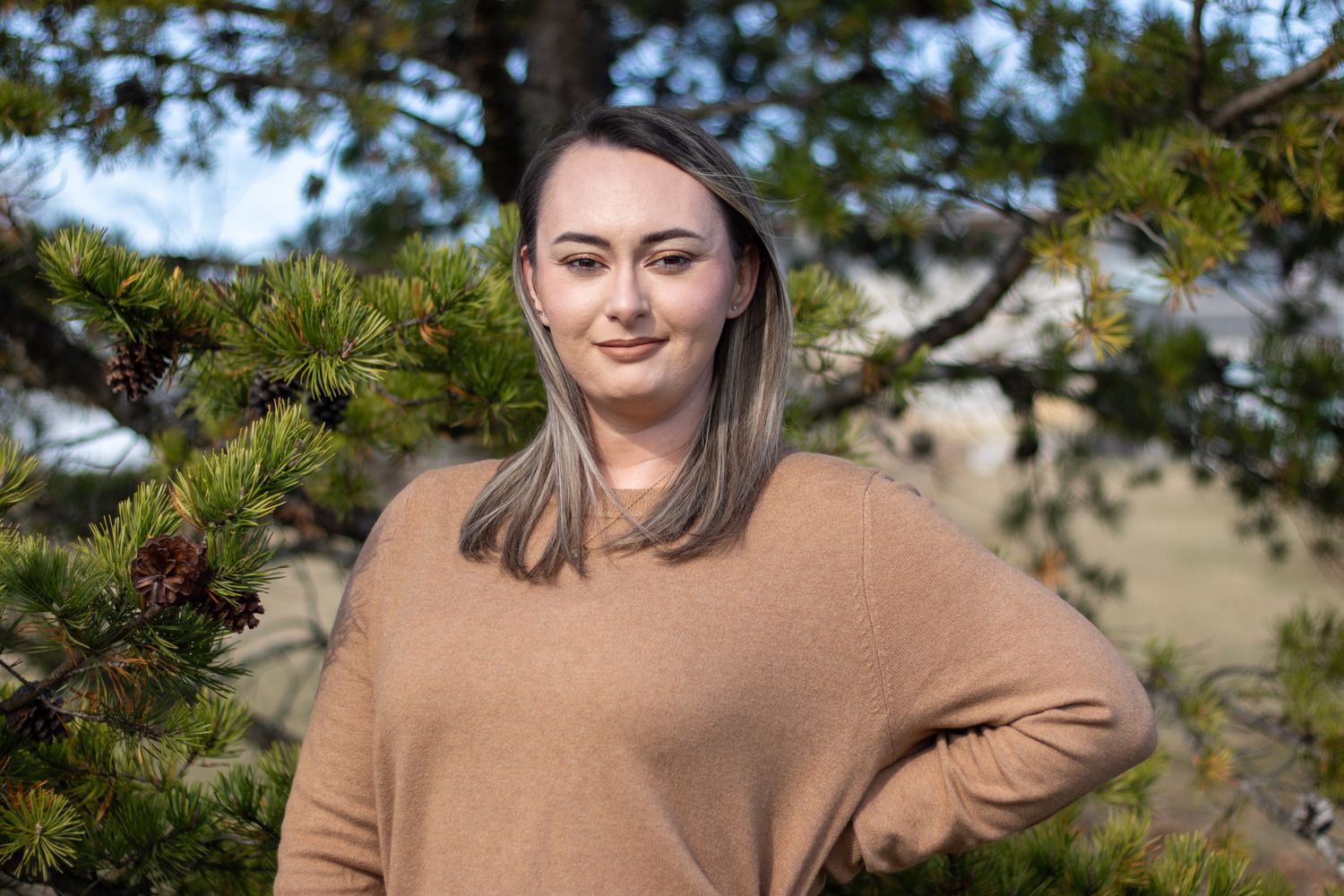 Woman smiling at the camera with her hand on her hip in front of the fall trees.