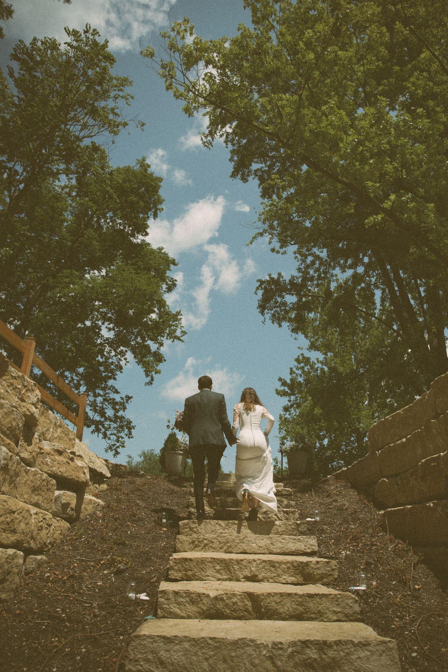 Couple ascends stone steps surrounded by trees on wedding day.