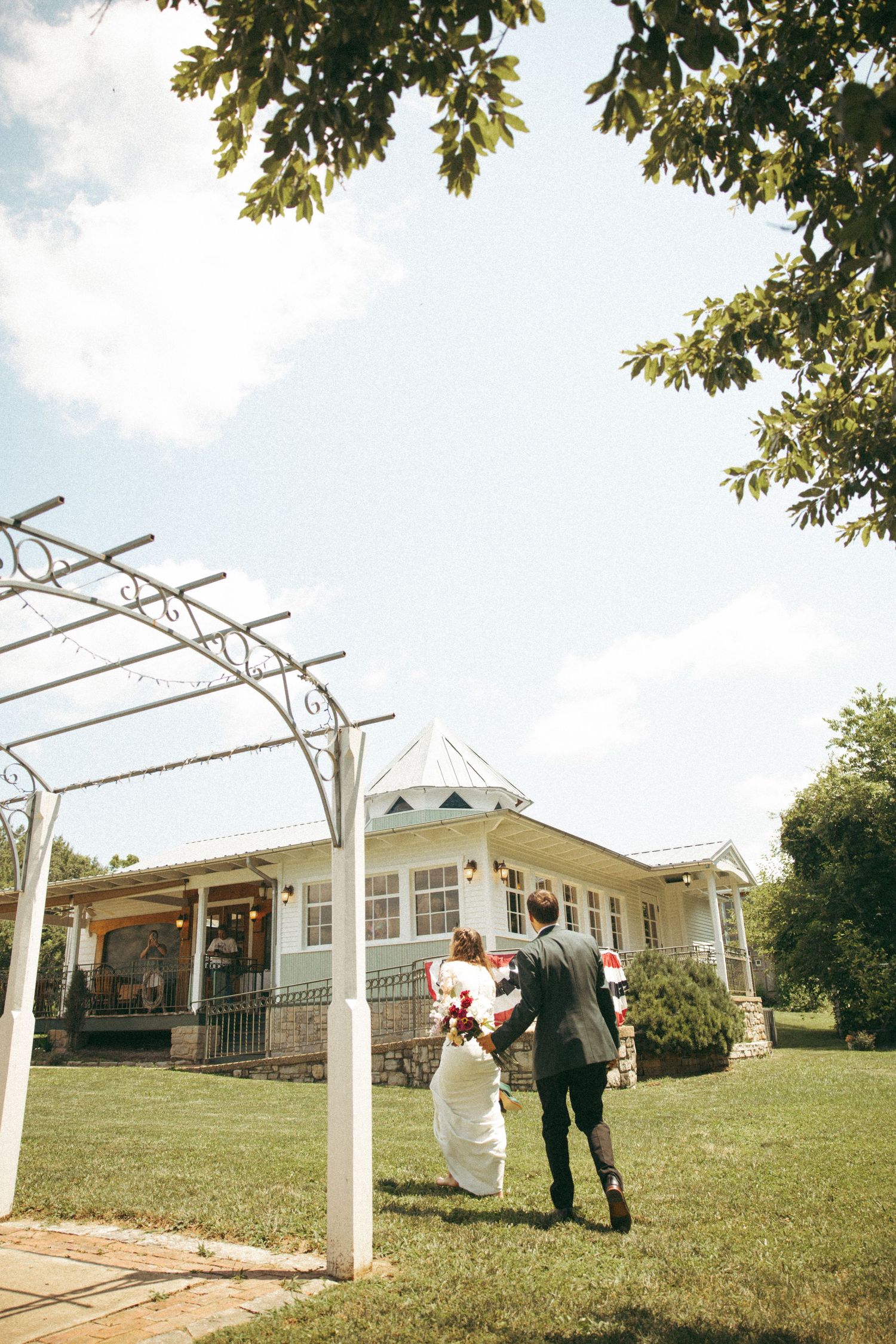 Newlyweds walk hand in hand toward white house under pergola on sunny day.