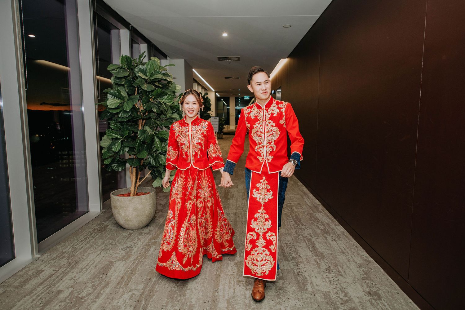 A couple walks down a hallway wearing matching red and gold traditional Chinese wedding outfits.