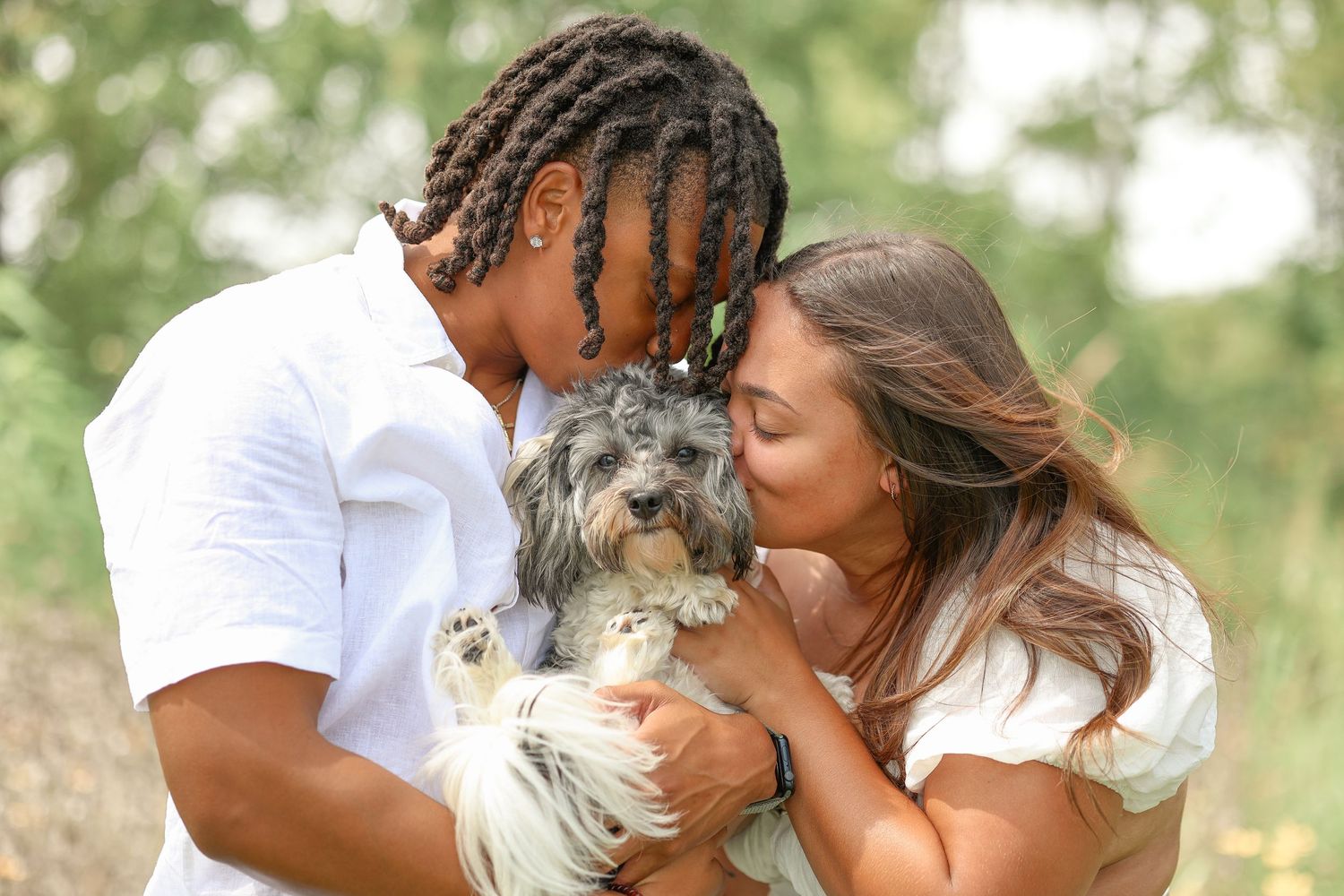 Couple in white shirts sharing a tender moment with their small gray dog outdoors.