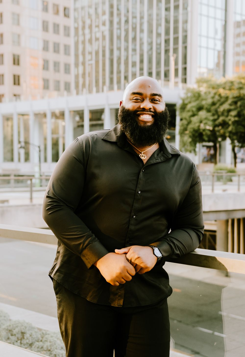 Professional portrait of someone smiling in a black button-up shirt against an urban backdrop of office buildings.