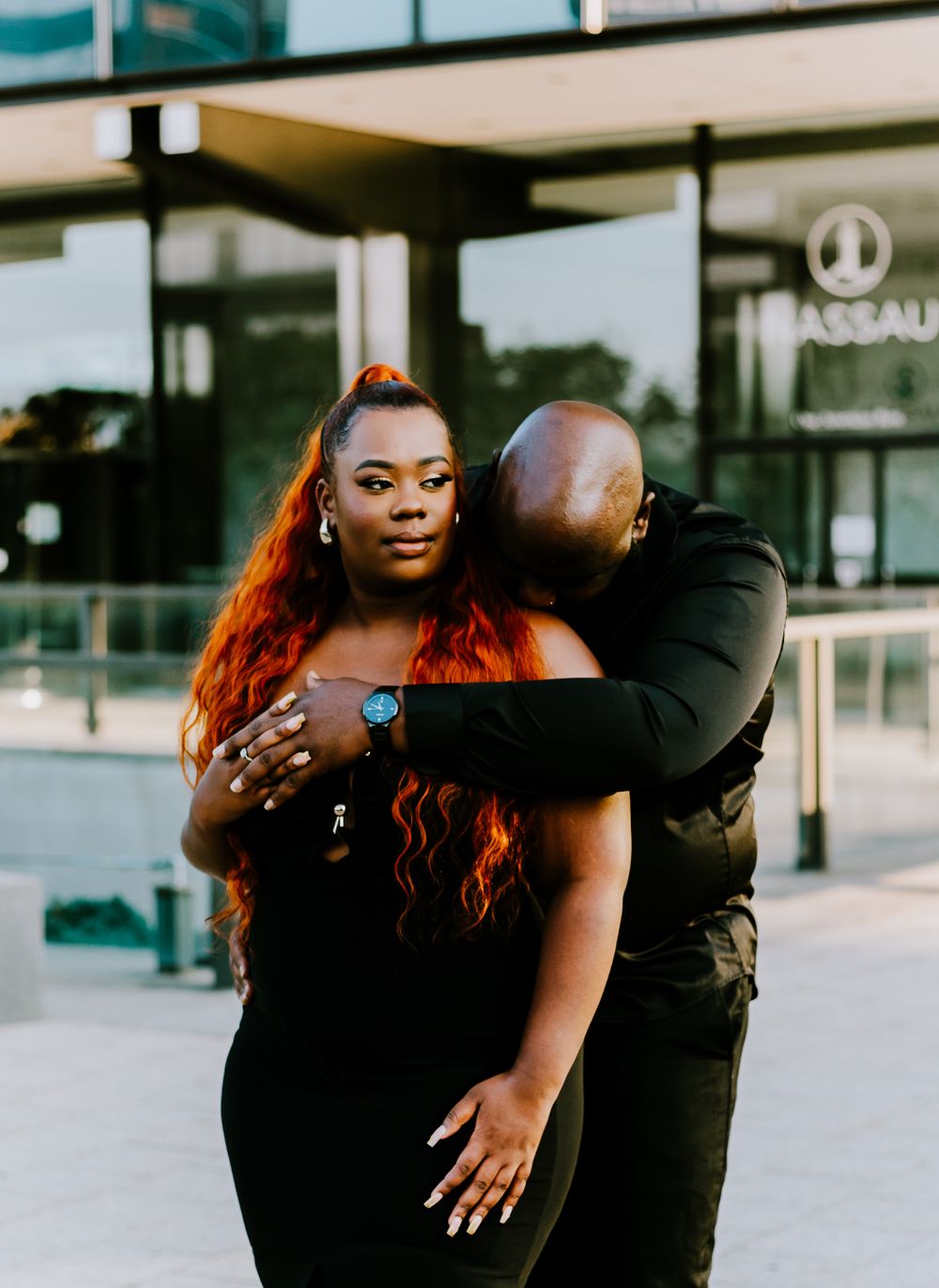 Romantic couple embracing outside a modern building as sunlight catches her flowing red hair.