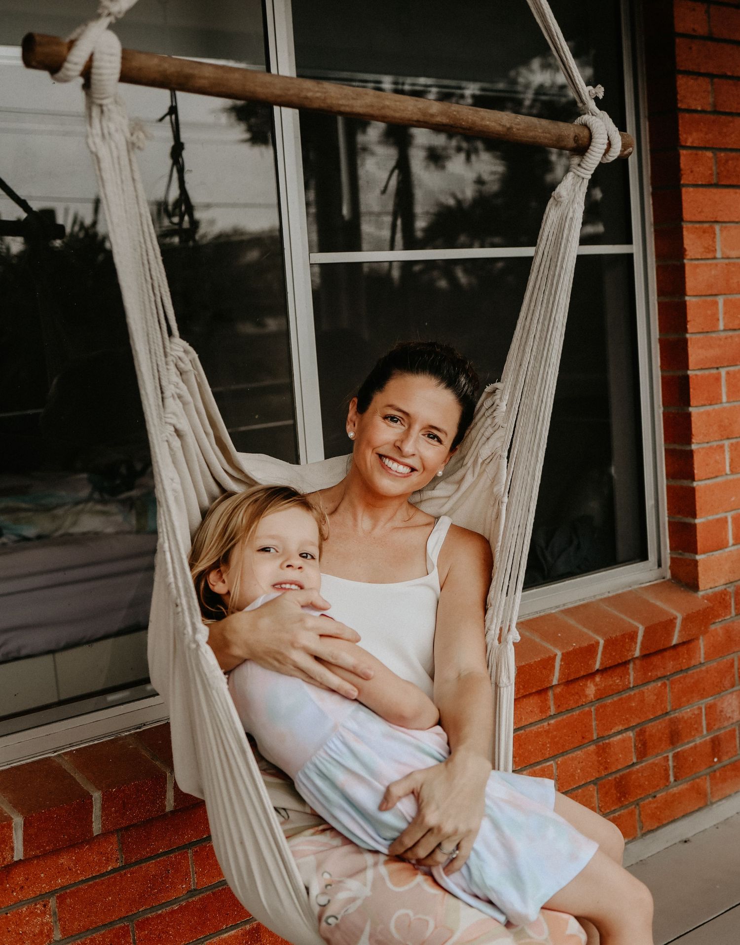 A relaxing moment shared in a white macrame hammock swing mounted outside a brick home with windows.