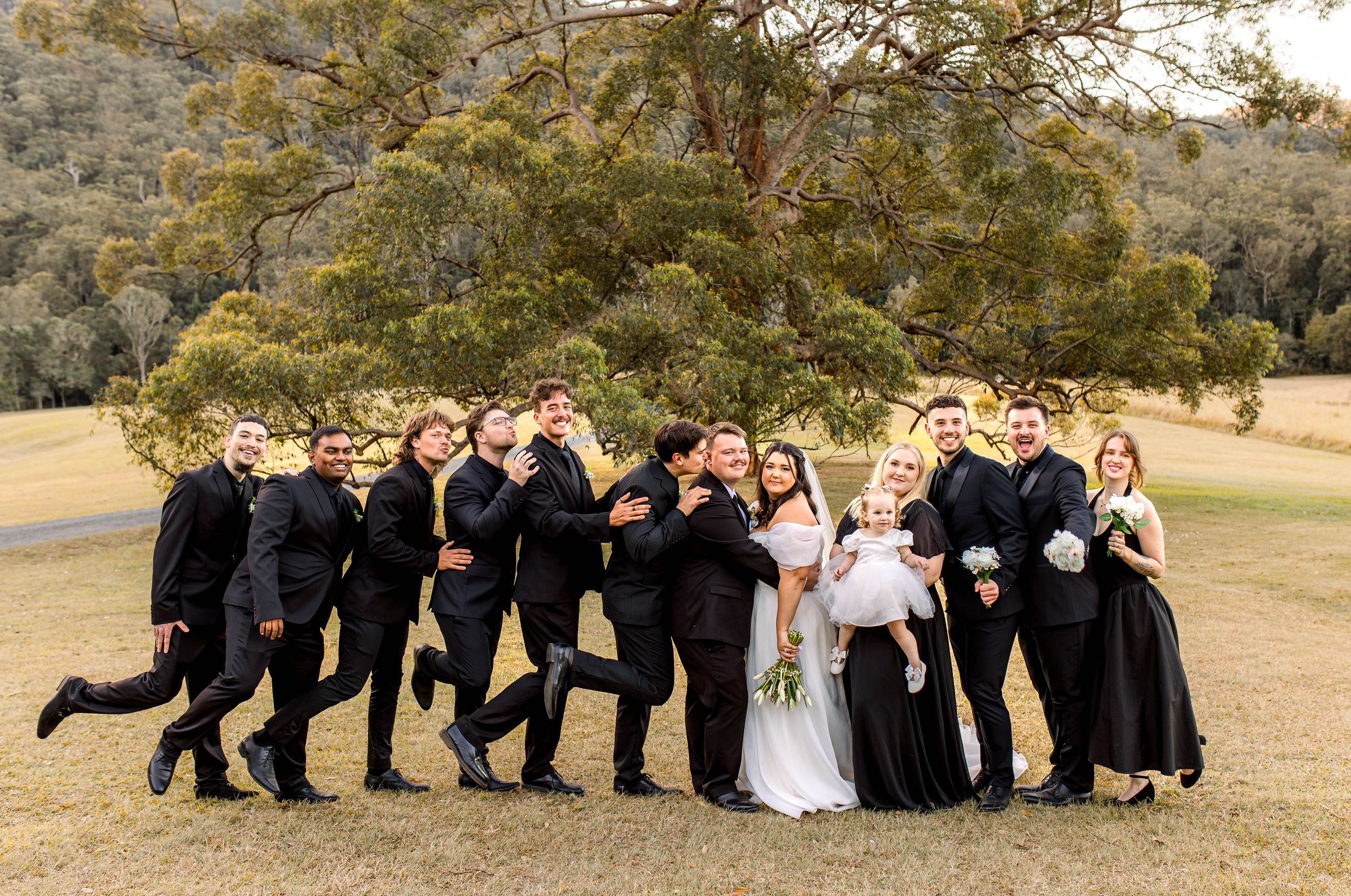 Wedding party in black attire posed creatively on a grassy hill at sunset.