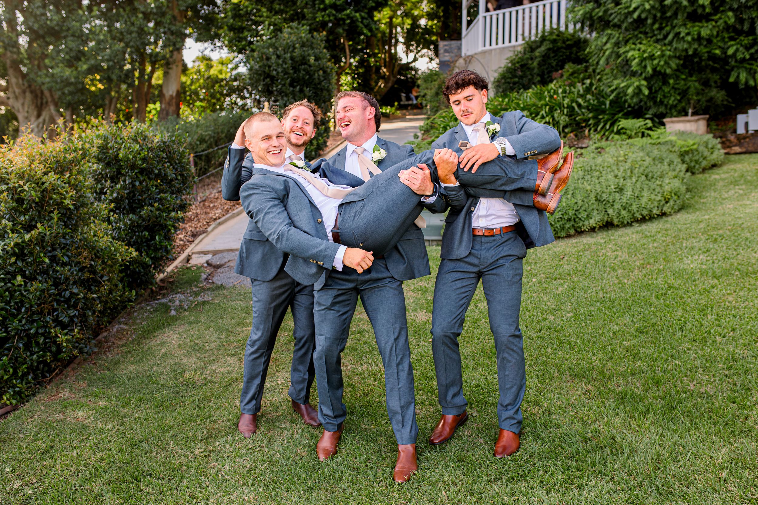 Wedding party members playfully jump and pose on a grassy lawn.