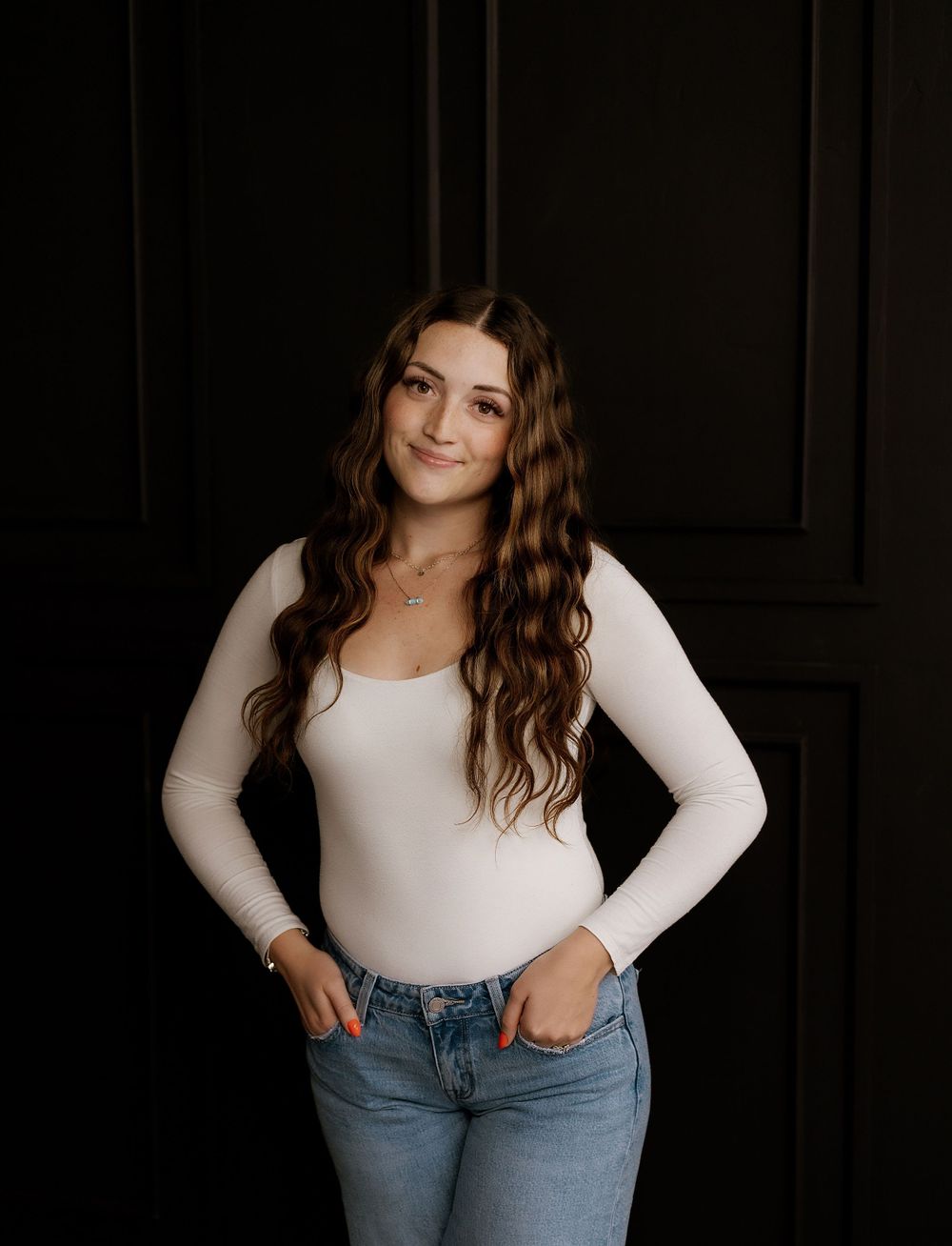 Casual portrait in white long sleeve top and jeans against dark background.