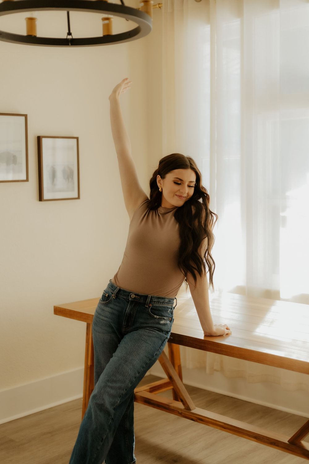 Dancer in a brown top and jeans practices poses in front of a window with natural lighting. Western