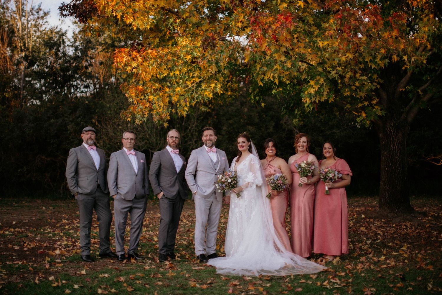 Wedding party poses together in front of autumn trees with golden fall foliage and scattered leaves on the ground.