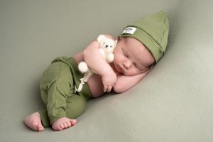 Sleeping newborn wearing olive green outfit and hat while cuddling small teddy bear.