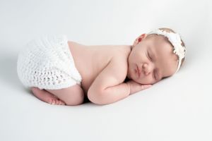 Sleeping newborn in white lace diaper cover wearing delicate floral headband on white background.