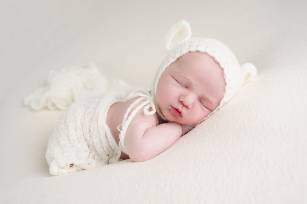 Sleeping newborn in white knit bear bonnet and wrap on cream background.