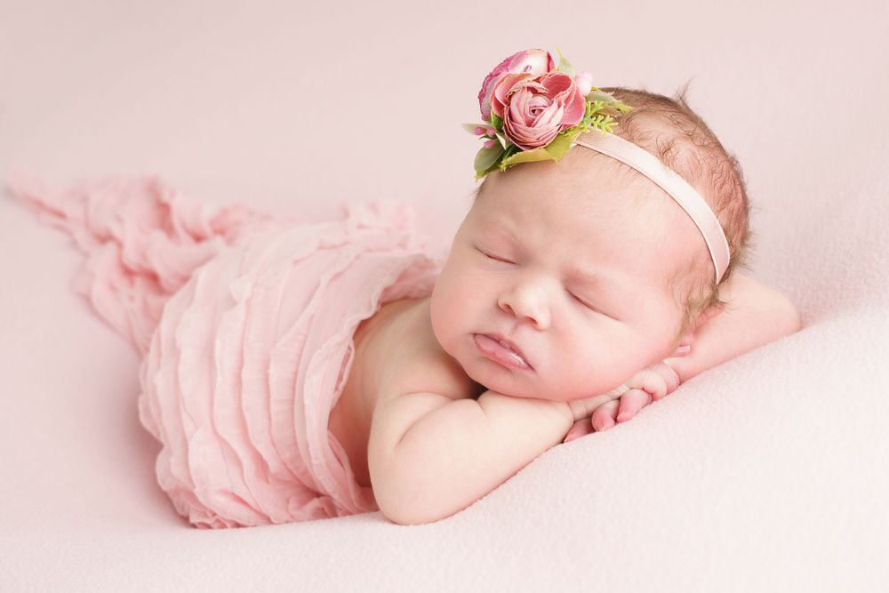 Newborn baby wearing pink wrap and floral headband sleeping peacefully in portrait.