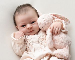 Newborn baby in cream outfit cuddles a soft pink plush bunny toy while lying on a white blanket.