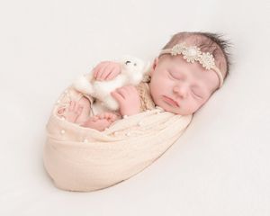 Sleeping newborn baby swaddled in cream wrap with lace headband, holding a white stuffed bear toy.