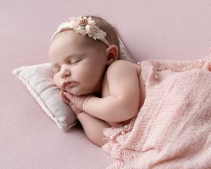Newborn baby girl sleeping peacefully on a pink pillow, wrapped in a soft pink blanket, wearing a floral headband.
