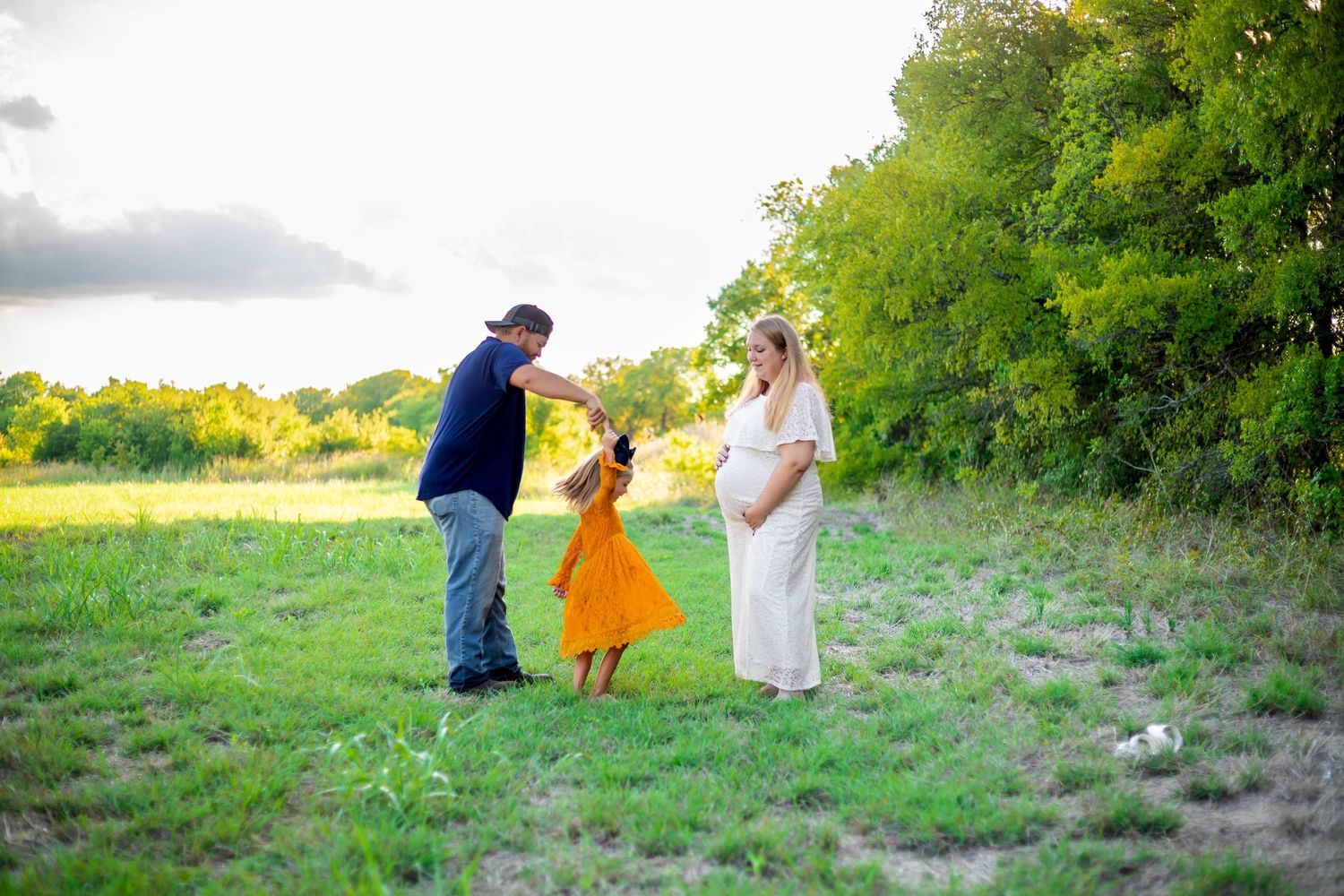 Family of three poses in sunny field during maternity photoshoot.
