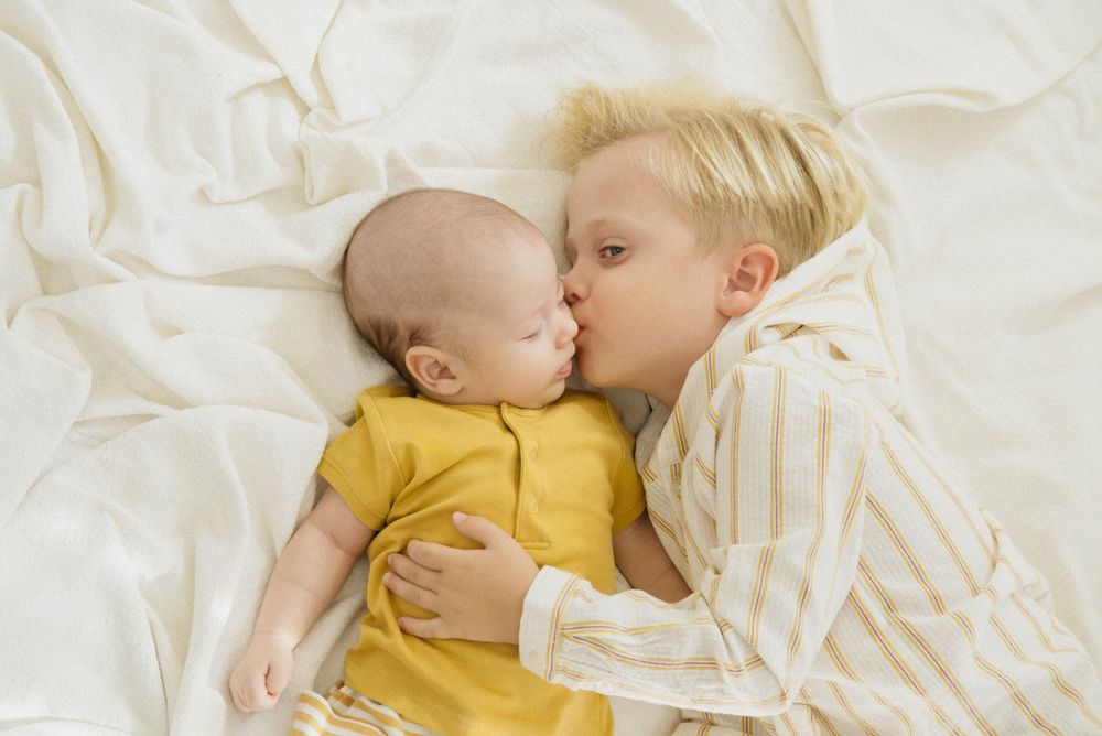 Dulce momento entre hermanos: un niño mayor con rayas besando a un bebé con un mono amarillo sobre ropa de cama blanca.