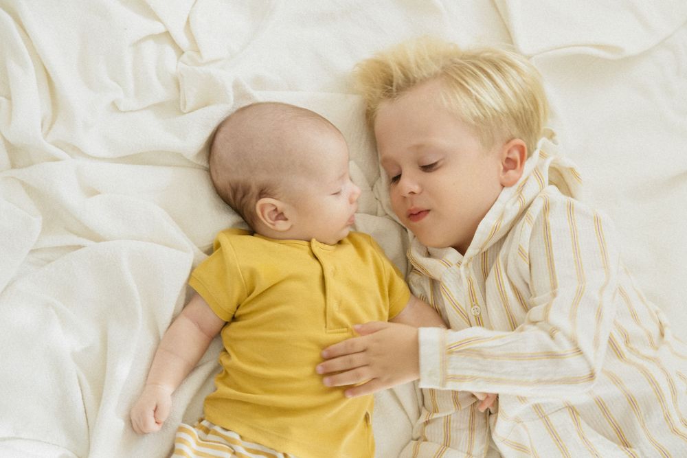 Retrato íntimo de hermanos que muestra un suave abrazo entre un bebé en amarillo y un niño mayor en sábanas blancas.