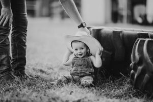 Black and white photo of a small child sitting in grass near tires while being touched on head by an adult hand.