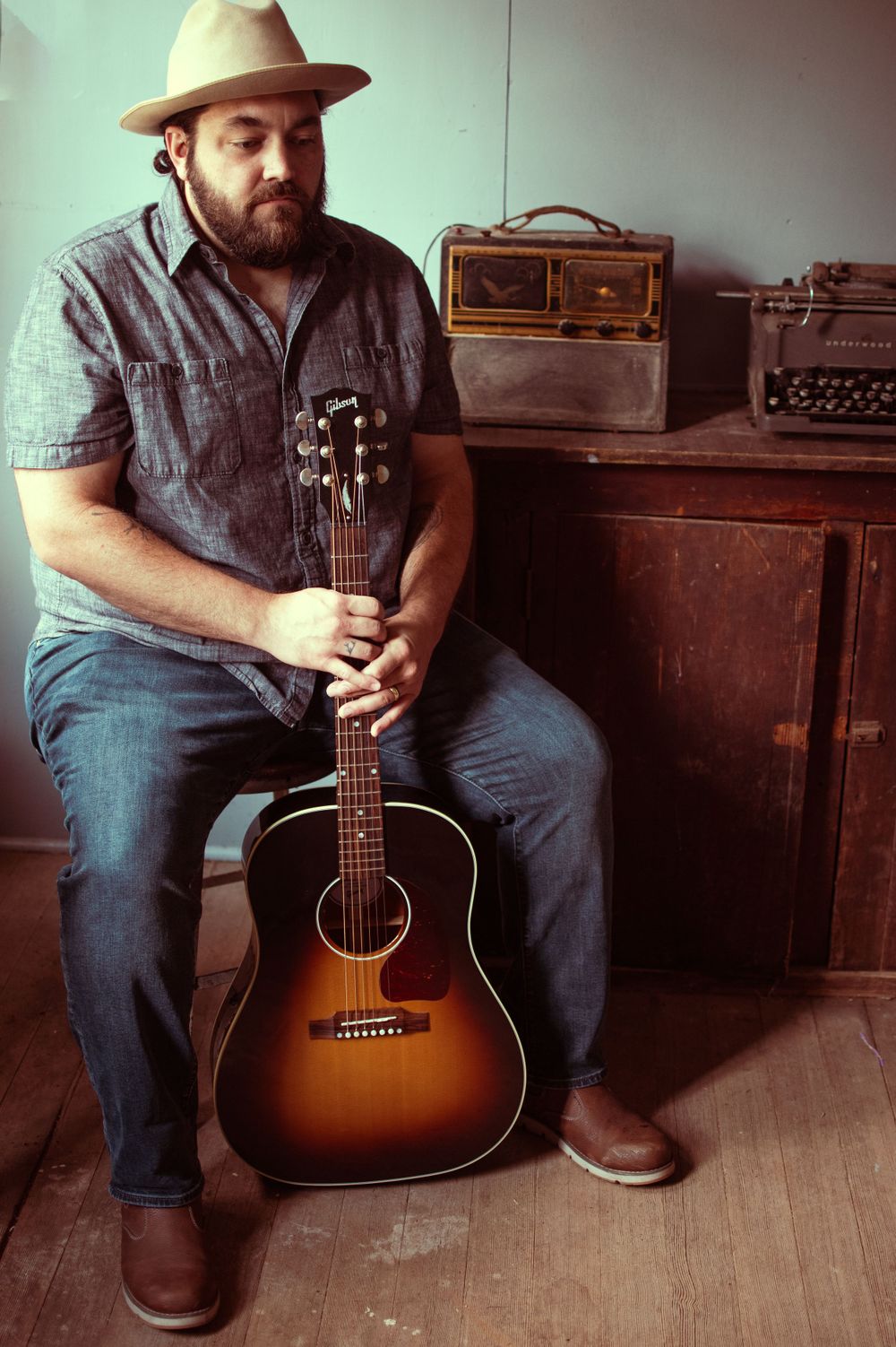 A musician in denim and hat sits with an acoustic guitar beside vintage radio and typewriter.