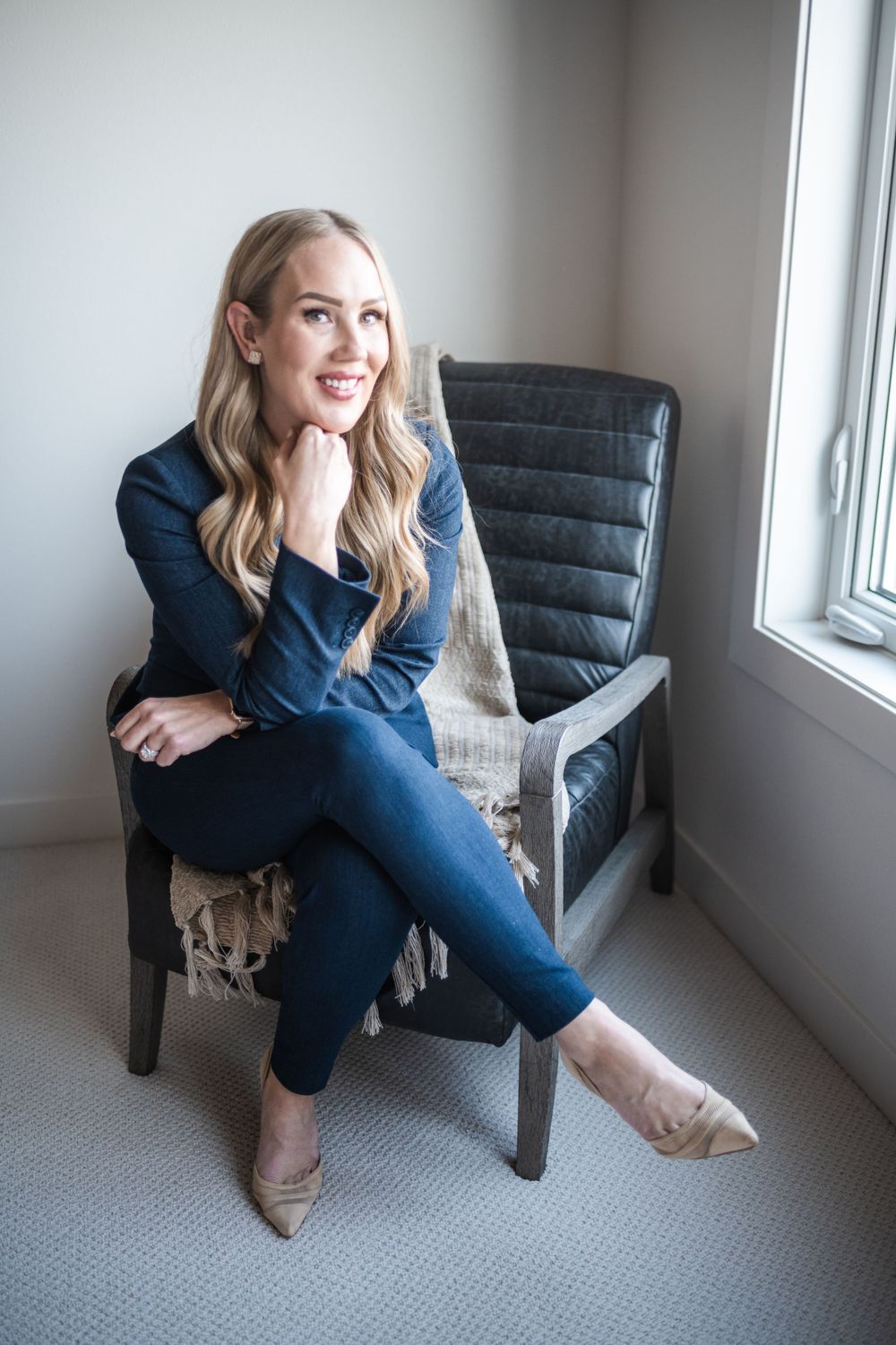 Woman in business attire sitting cross-legged on grey chair by window in professional setting.