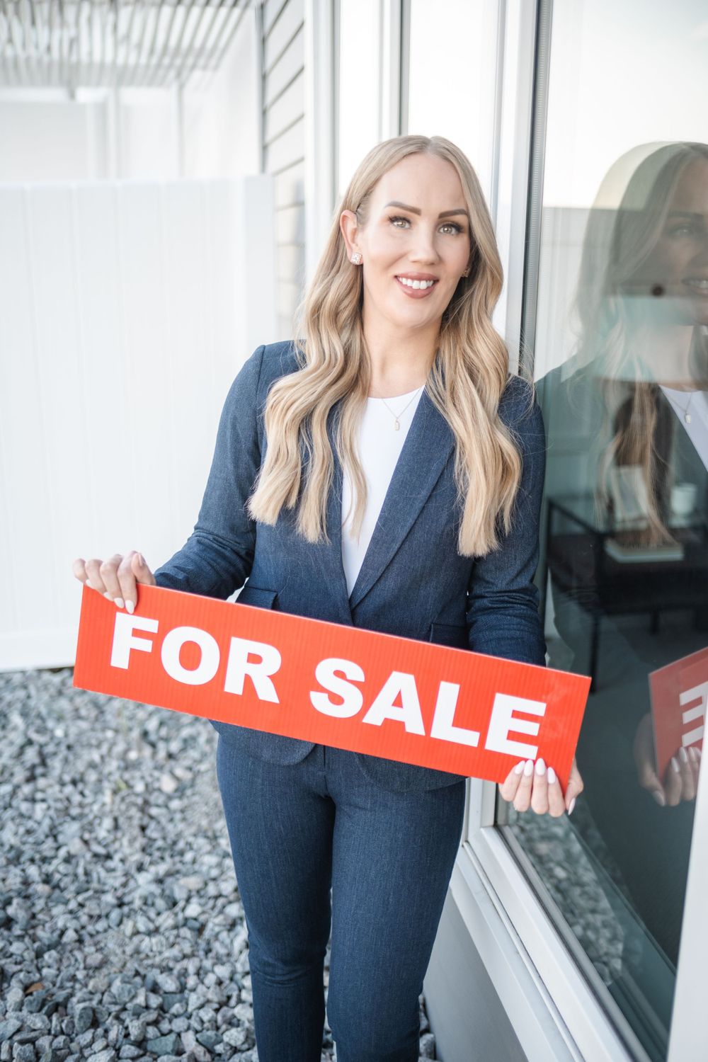 Realtor in navy suit holding red For Sale sign while standing by window in professional setting.