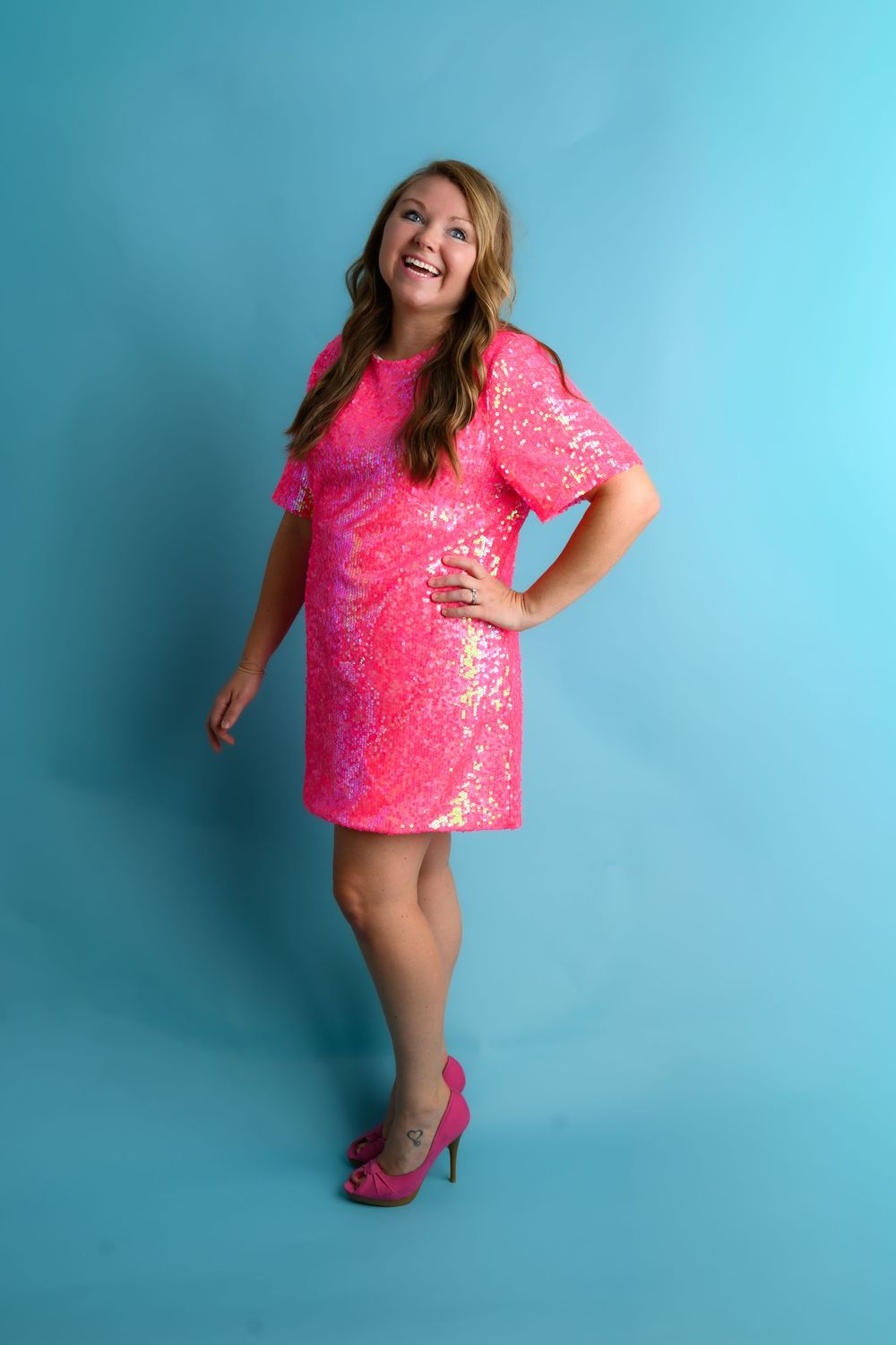 A person in a bright pink sequined dress and matching heels poses playfully against a turquoise blue background.