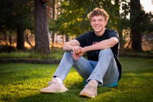 Casual outdoor senior portrait on grass with golden evening sunlight filtering through trees.