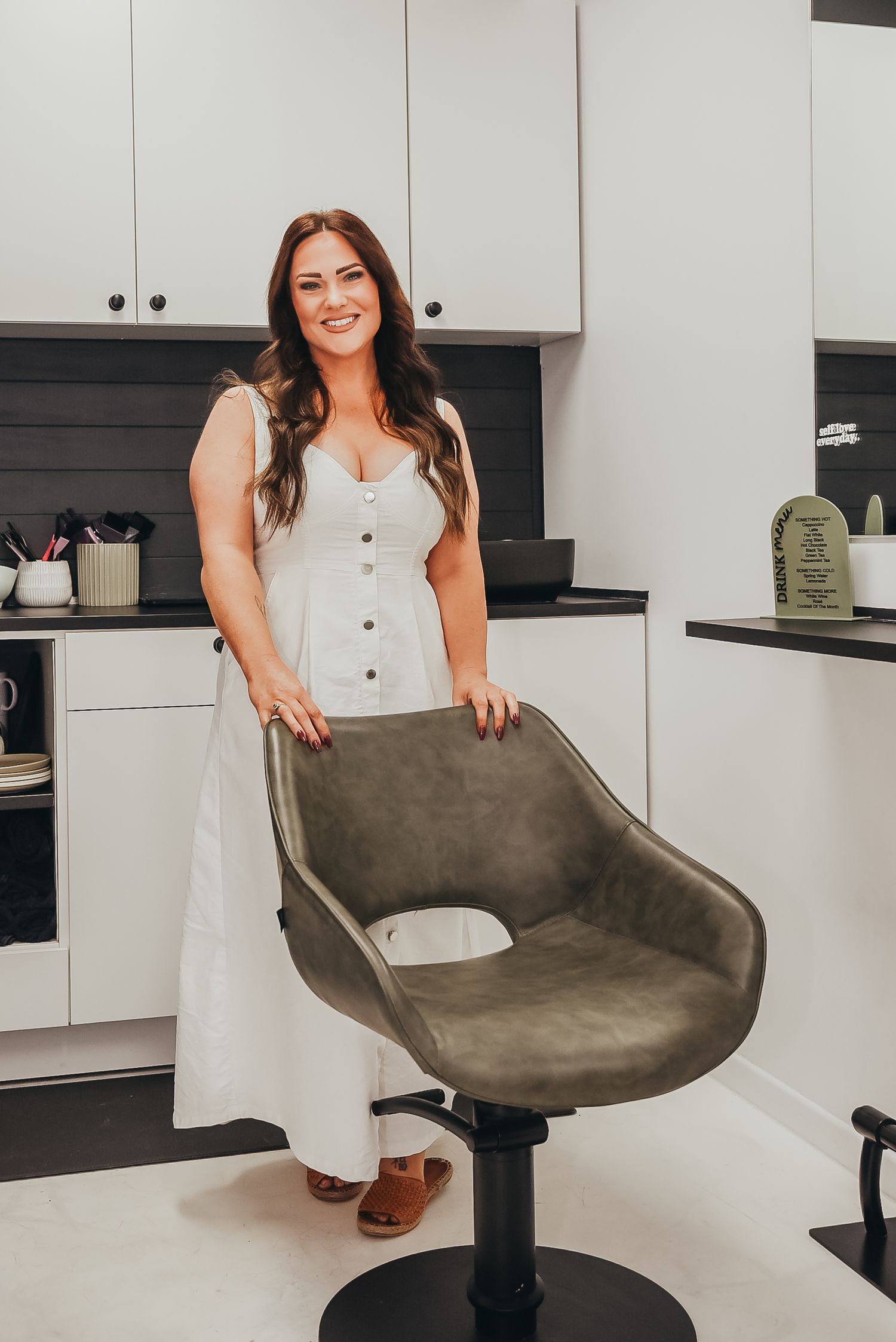Hair stylist in white dress standing next to brown salon chair in modern white kitchen.
