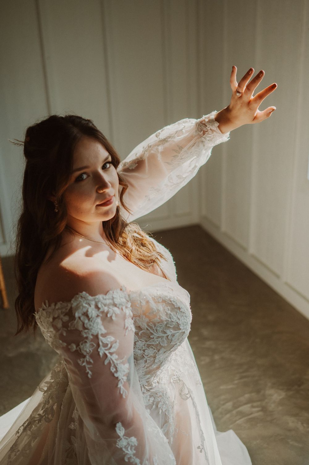 A bride in a long-sleeved lace wedding dress poses gracefully in a moody indoor setting with natural light.