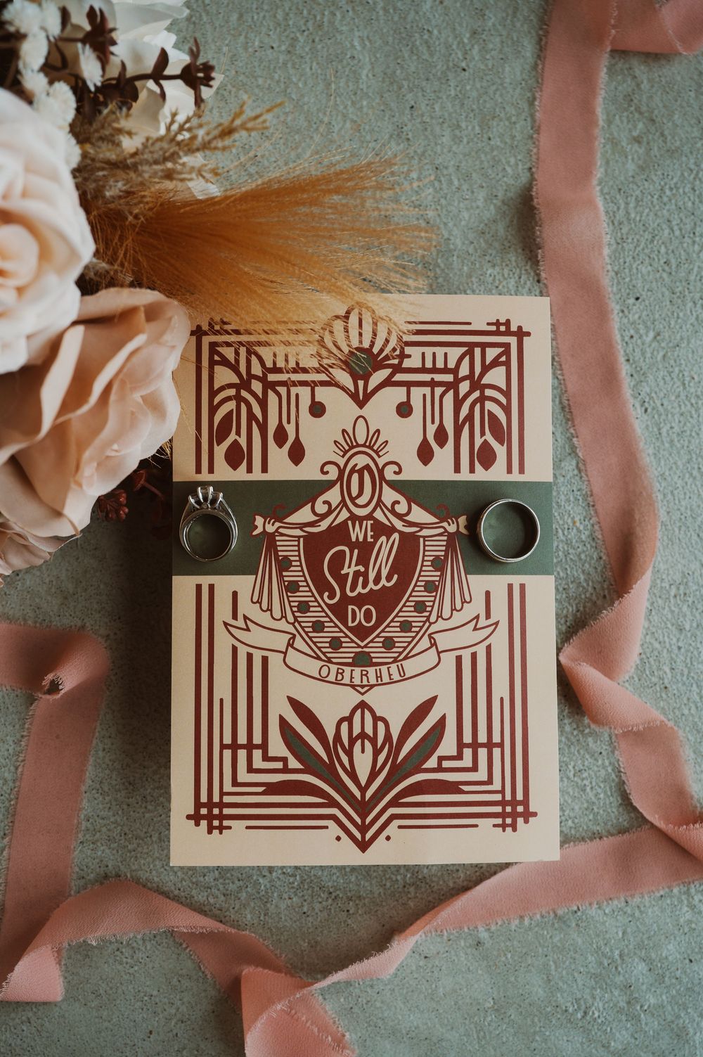 Art deco wedding invitation reading We Still Do with two gold rings and a pink floral bouquet on a teal surface.