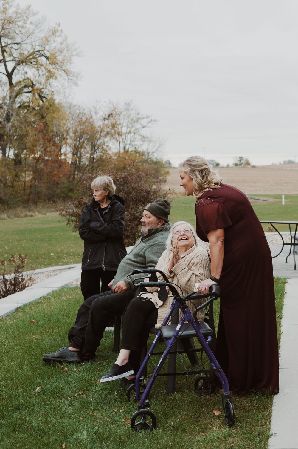 Elderly people gathered outdoors on grass, one in wheelchair with walker nearby, assisted by woman in burgundy dress.