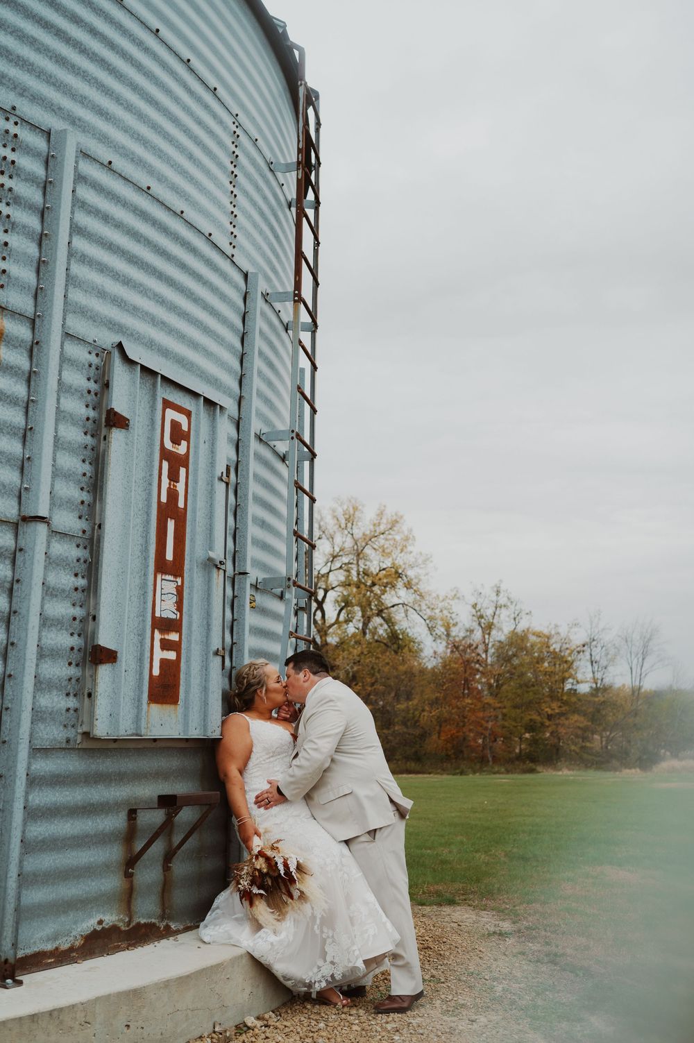 Bride and groom kissing beside a rustic grain silo at an outdoor fall wedding venue.