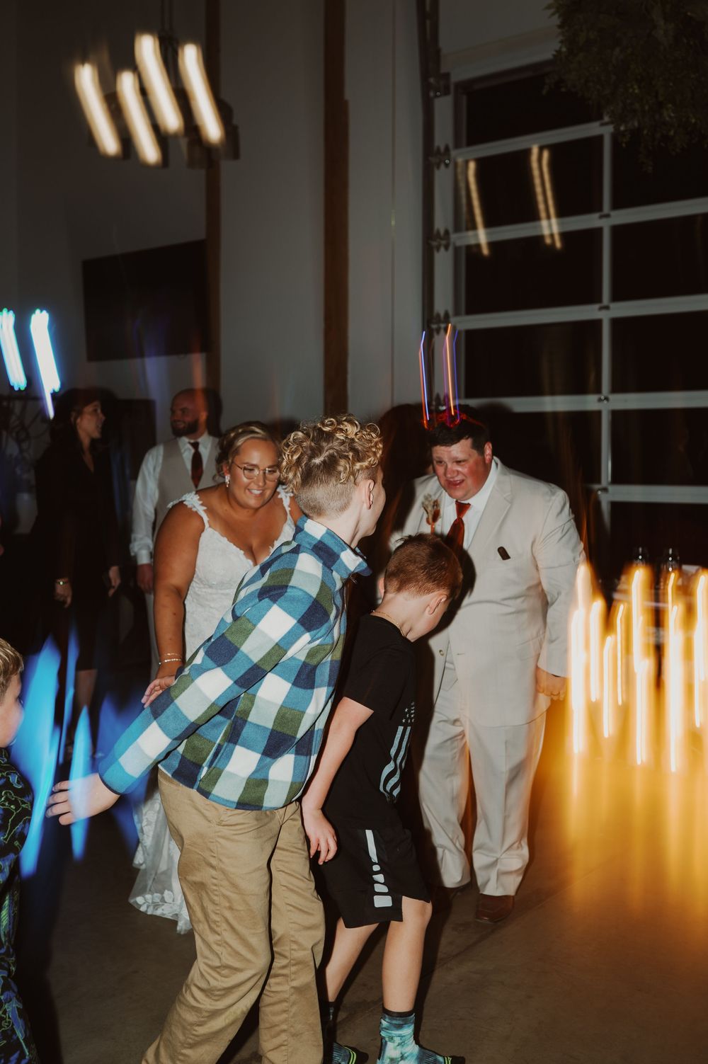 Bride and groom dance with children on colorful light-filled dance floor at lively wedding reception.