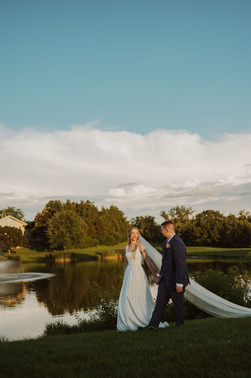 Couple walks along serene lakeside during golden hour with bride's veil flowing in the breeze.