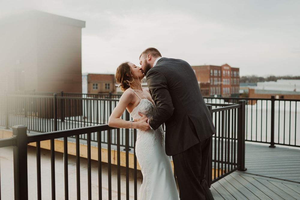 A couple shares a romantic kiss on a waterfront balcony during their wedding celebration at sunset.
