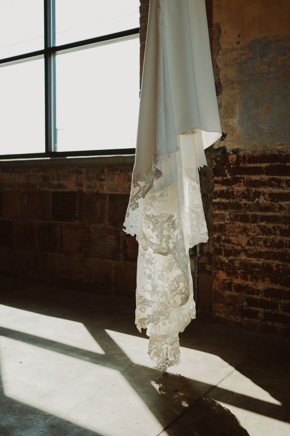 White lace wedding dress hanging by a window casting dramatic shadows on exposed brick walls in an industrial space.