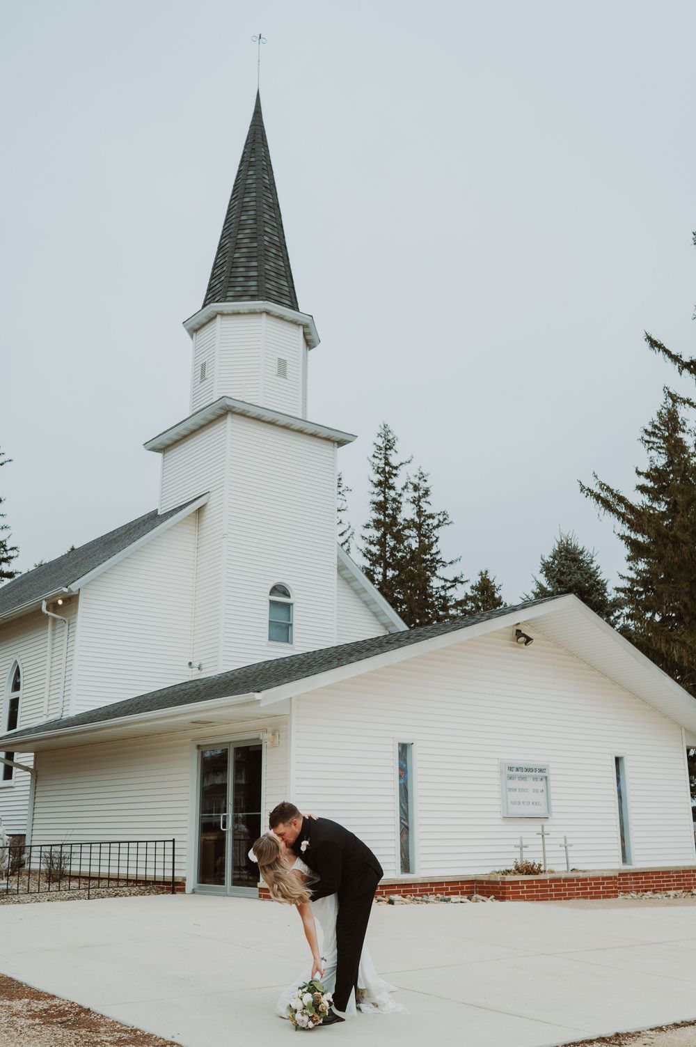 Bride and groom share a romantic dip kiss in front of a white church with a steeple on their wedding day.