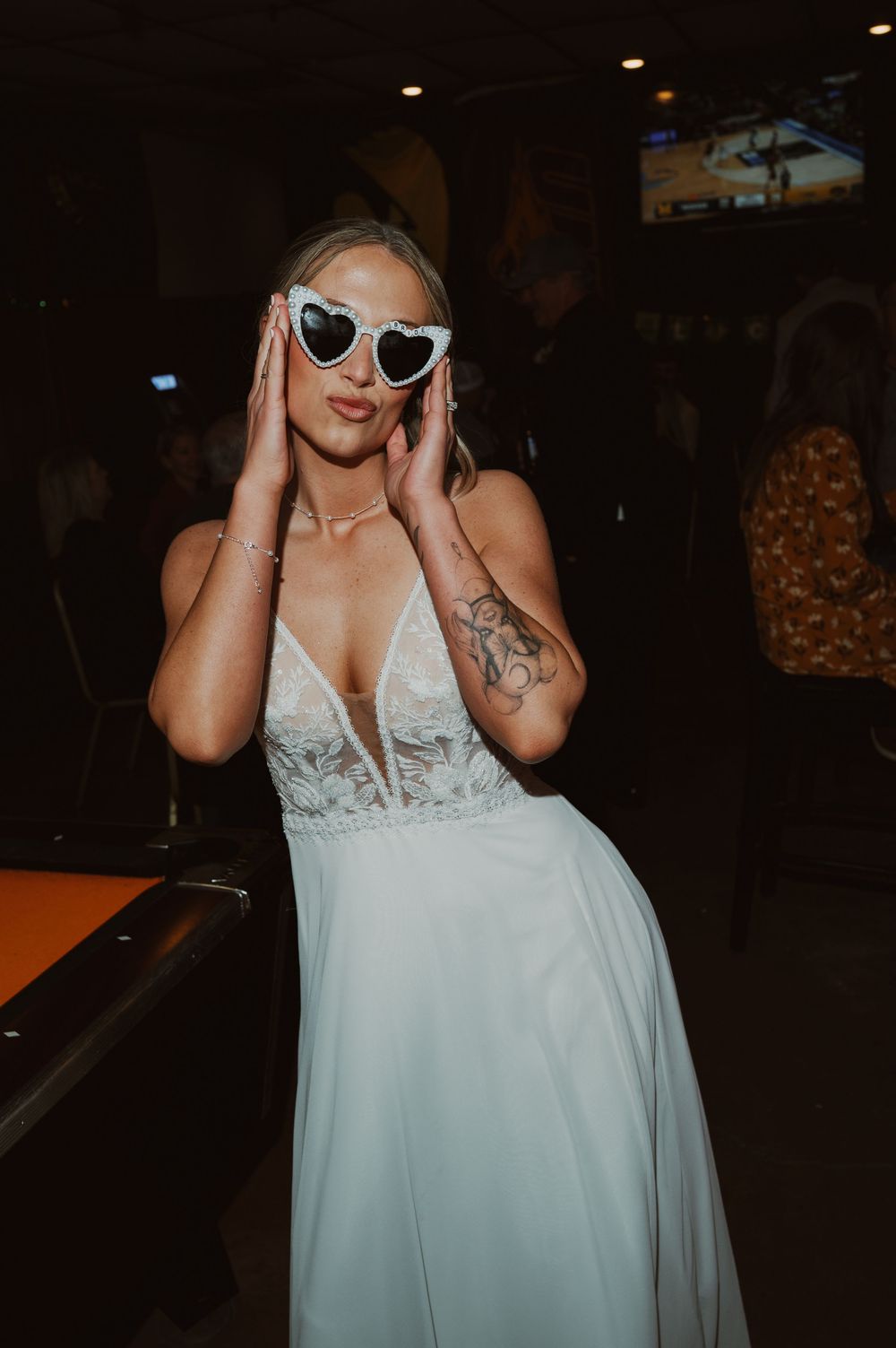 Bride in white lace gown wearing heart-shaped sunglasses poses at wedding reception venue.