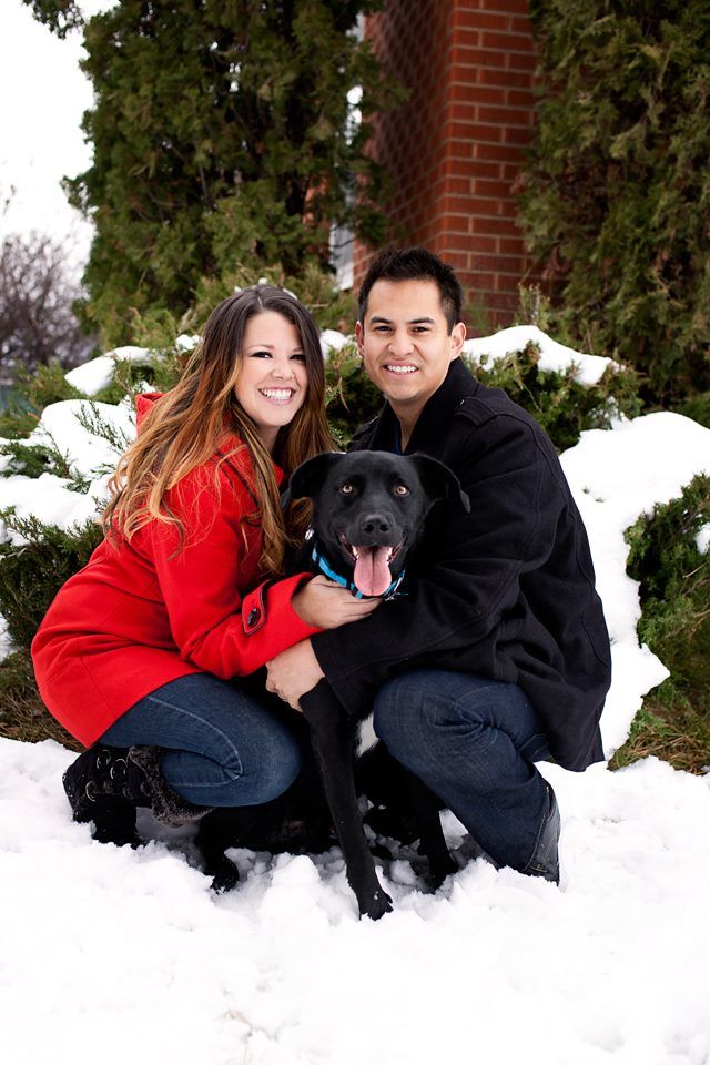 A couple in winter attire poses with their black Labrador dog in the snow near a brick building.