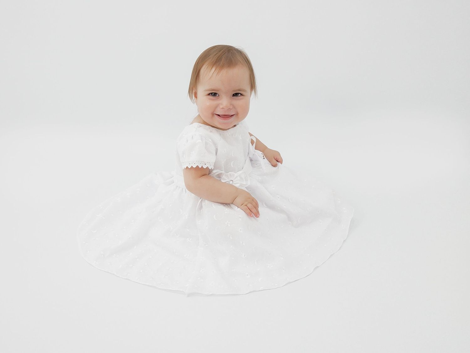 Baby in a white dress sits on a light backdrop during a minimalist studio photo session.
