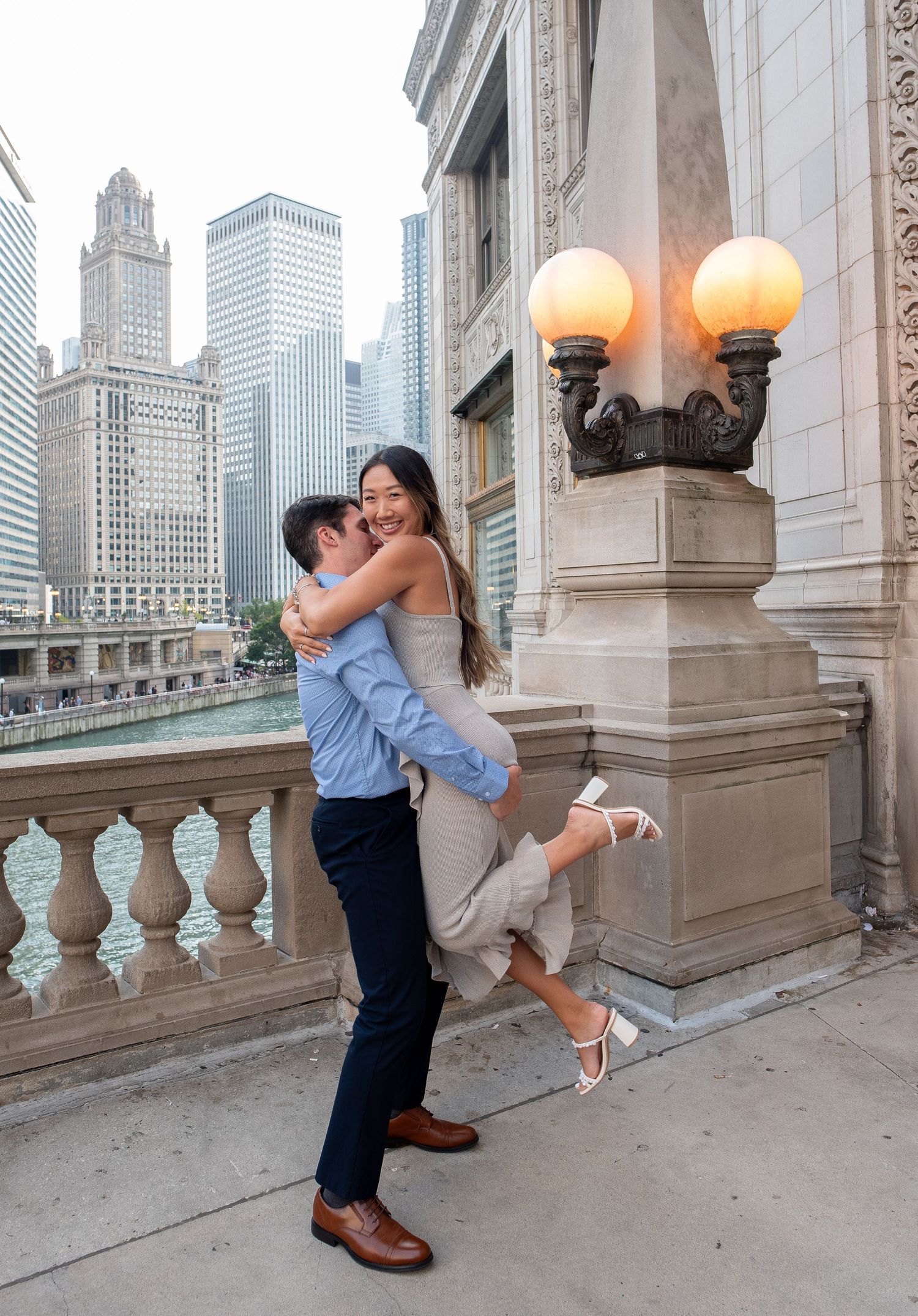 A romantic couple embraces and kisses on an elegant stone balcony with the Chicago skyline visible in the background.