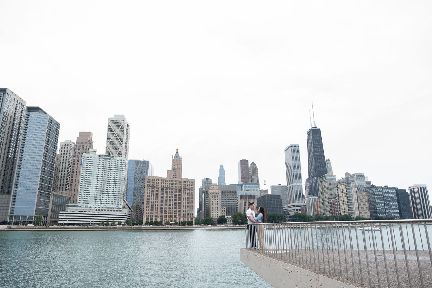 A couple stands on the Chicago Riverwalk with the iconic downtown skyline and skyscrapers in the background.