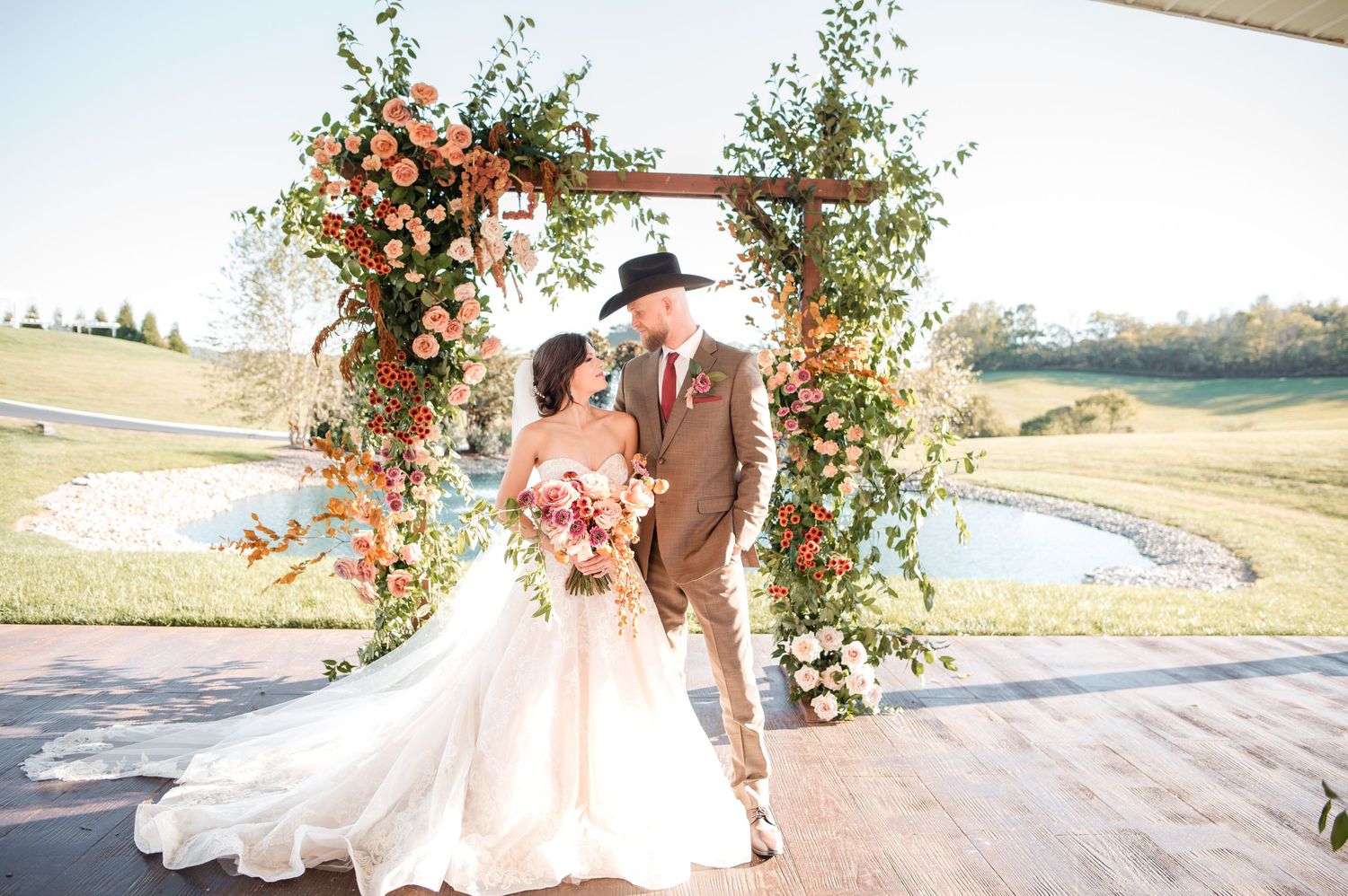 A romantic couple embraces during their wedding ceremony under a floral arch at an outdoor venue during sunset.