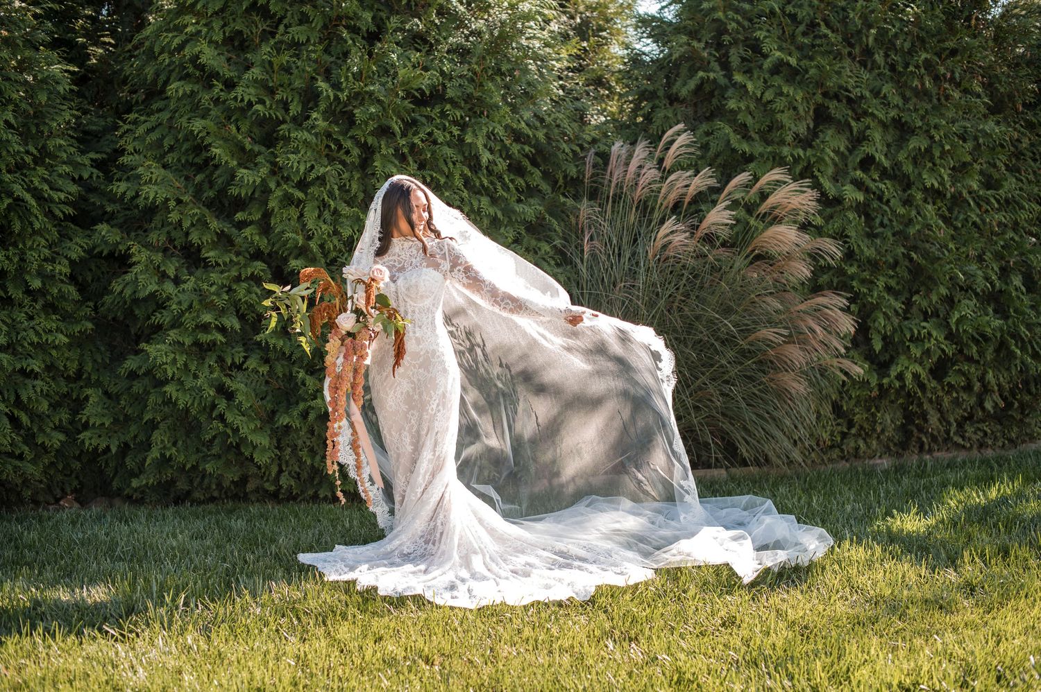A bride in a flowing white dress poses gracefully on a grassy hillside in a garden setting.