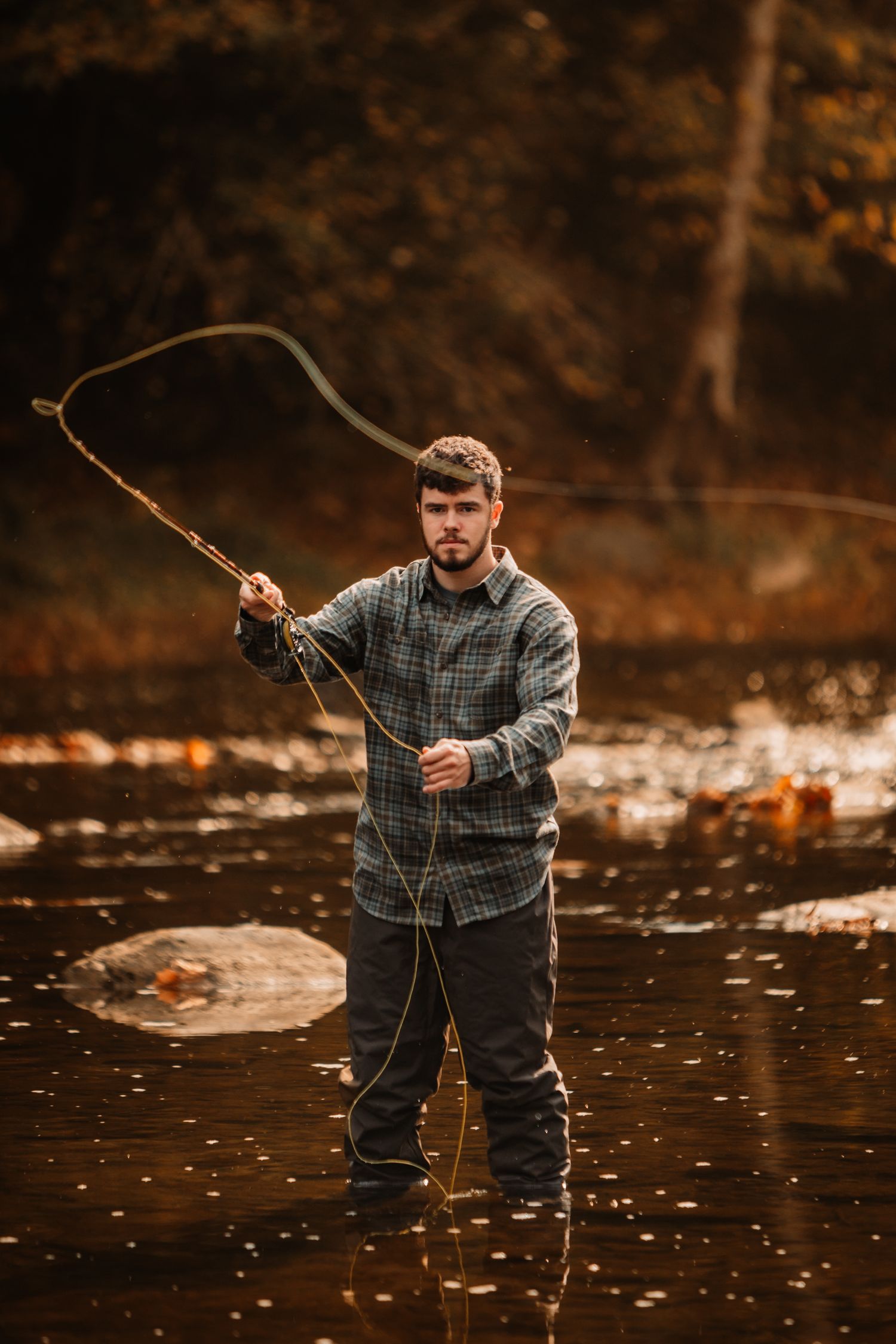 Person in plaid flannel fishes with a rod in a serene autumn stream.