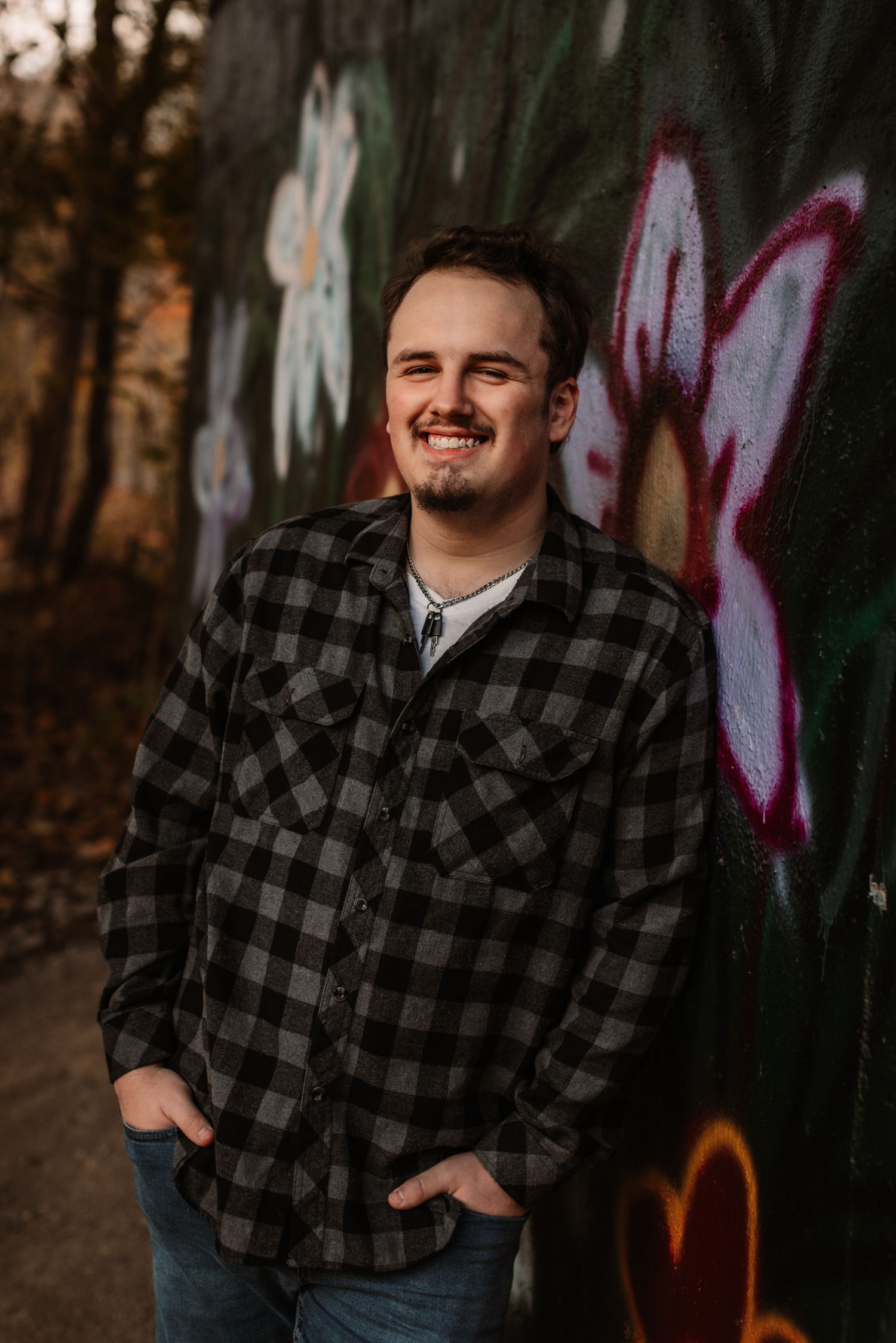 A person in a black and gray plaid flannel shirt poses outdoors against a graffiti wall at sunset.