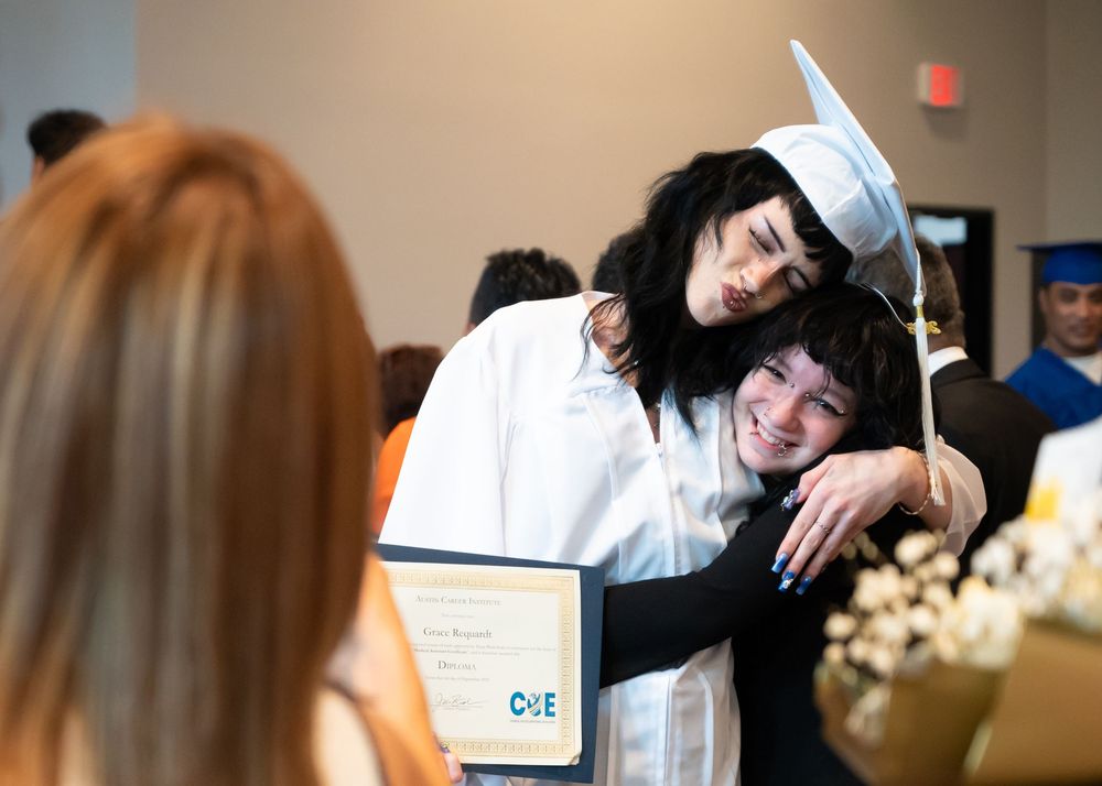 A graduate in white cap and gown shares an emotional hug while holding a diploma at graduation.